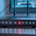 Roteador wifi sobre mesa escura com o LED de Internet aceso em vermelho entre os demais LEDs brancos, telas com codigo desfocadas ao fundo